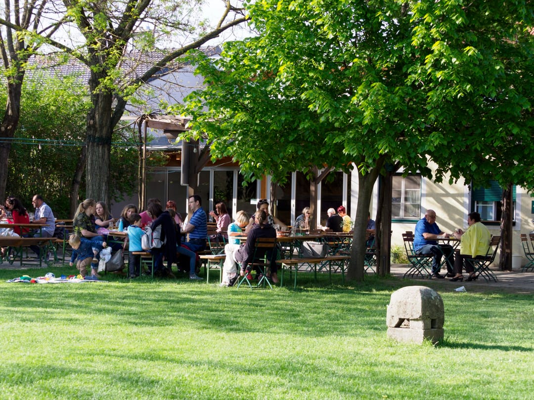 Gut besuchter Gastgarten mit Gästen an Tischen unter Bäumen im Frühling