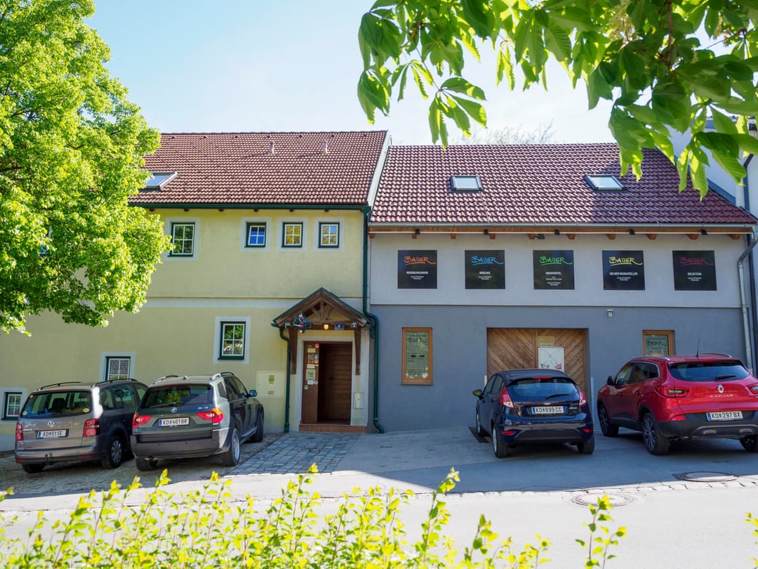 Außenansicht des Buschenschank-Gebäudes mit Bauer-Beschilderung und Parkplatz
